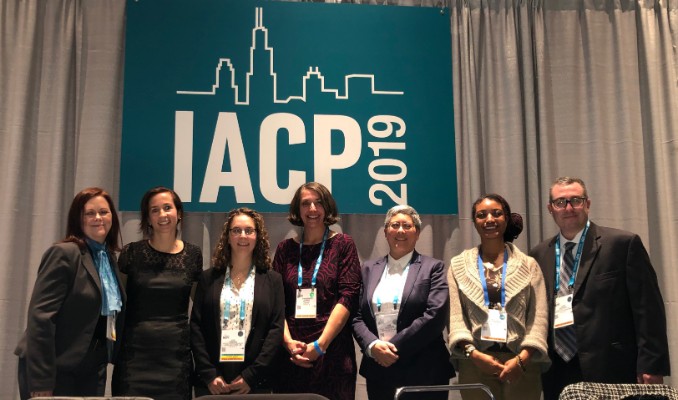 6 female and 1 male panelists all in a row posing with big smiles in front of the IACP 2019 banner.